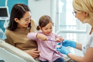 تُظهر الصورة تجربة تعليمية ممتعة خلال **أول زيارة لطفلك في عيادة الأسنان**، حيث تجلس طفلة صغيرة في حضن أمها على كرسي الطبيب.
تقوم طبيبة الأسنان بمساعدة الطفلة وتدريبها على استخدام الفرشاة لتنظيف مجسم تعليمي للأسنان بطريقة تفاعلية.
يعكس المشهد البيئة الآمنة والمشجعة التي تساعد الأطفال على كسر حاجز الخوف والاعتياد على العناية بصحة الفم والأسنان.